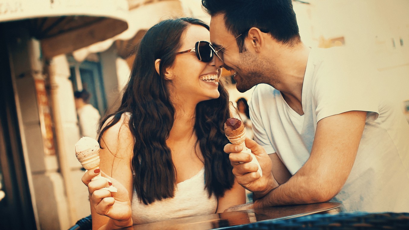 The couple is smiling while eating ice cream, getting closer together to kiss.